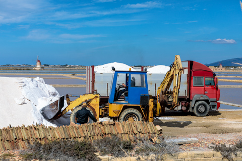 Heavy machinery loads large pile of sea salt at the Saline Genna salt pans near Marsala, Sicily, Italy, with shallow salt flats stretching into the distance.