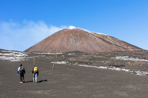 Two hikers walk across a dark volcanic plain toward Mount Etna in winter, with patches of snow scattered across the ground beneath a clear blue sky.