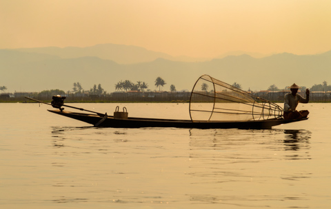 An Intha fisherman sits in a narrow wooden boat on Inle Lake, holding a conical fishing net as calm water reflects the warm light of early morning in Myanmar.
