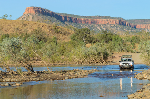 A four-wheel-drive vehicle crosses the shallow Gibb River beneath red escarpments along the Gibb River Road in Western Australia.