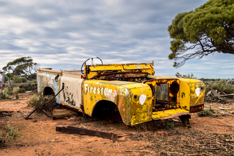 The rusted remains of a vintage Land Rover sit abandoned near Koonalda Homestead on the Nullarbor Plain in South Australia, surrounded by red earth, low scrub, and wind-shaped trees.