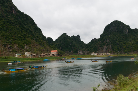 Small wooden boats travel along the Sông Son River in the Phong Nha region of Vietnam, with steep limestone karst hills rising above riverside villages under an overcast sky.