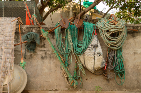 Anchors, coiled fishing ropes, and woven nets hang against a concrete wall in a coastal fishing village near Bãi Xép, Vietnam, reflecting the tools of daily maritime life.