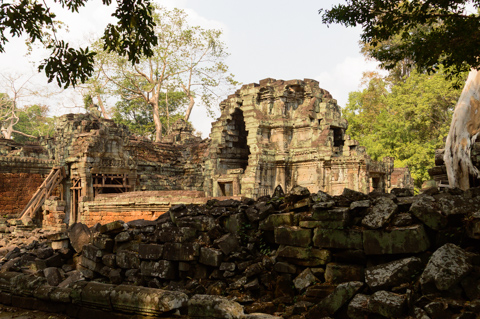 Collapsed stone walls and weathered towers of a Khmer temple stand among trees at Angkor Archaeological Park in Cambodia, with scattered blocks marking centuries of decay and restoration.