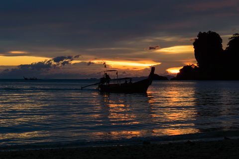 A long-tail boat floats near shore at sunset at Tonsai Beach in Thailand, silhouetted against fading light over the Andaman Sea and calm coastal waters.