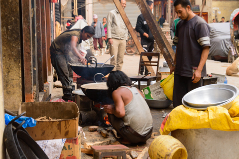 Street cooks prepare food over large woks along a roadside in Kathmandu, Nepal, as smoke rises from open fires and pedestrians move through the busy street.
