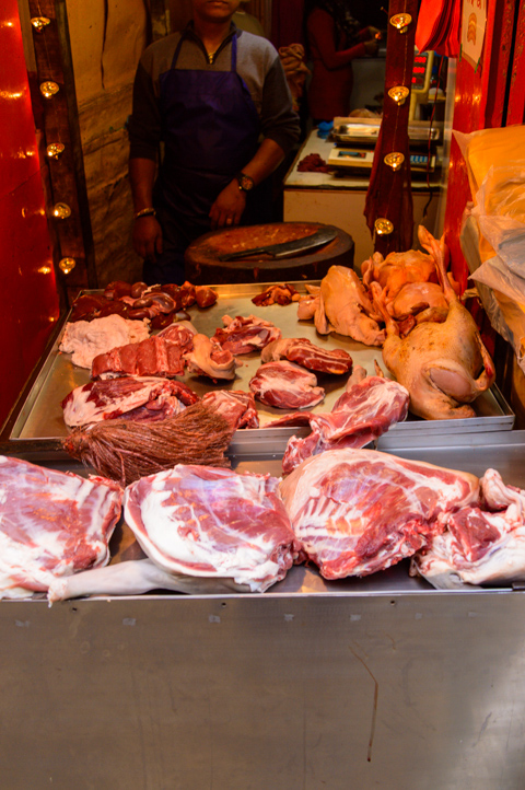 Cuts of raw meat and whole poultry are displayed on a metal counter inside a small neighborhood butcher shop in Kathmandu, Nepal, with the vendor standing behind the stall.
