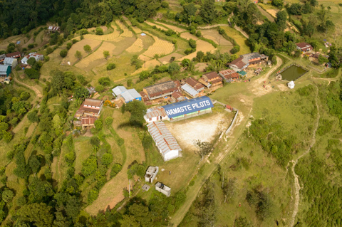 An aerial view of a rural village near Pokhara, Nepal, with terraced fields, scattered homes, and a large rooftop sign reading “Namaste Pilots” visible among the farmland.
