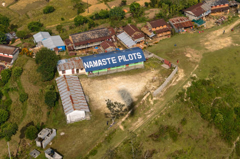 An aerial view of a village school near Pokhara, Nepal, with “Namaste Pilots” painted across the roof, acknowledging donations from visiting paragliding pilots.