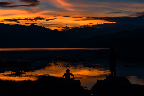 Two silhouetted men fish along the edge of Fewa Lake at sunset in Pokhara, Nepal, as orange and purple light reflects across the still water beneath dark mountain outlines.