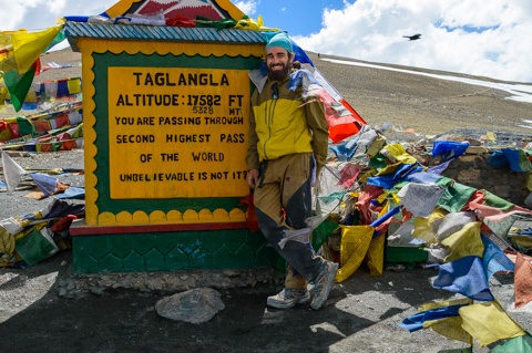 A traveler stands beside a sign marking Tanglang La Pass in the Indian Himalayas, surrounded by prayer flags at an altitude of 17,582 feet along a high mountain road.