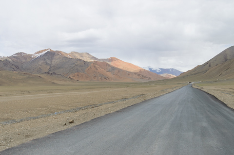 A long, straight paved road stretches across the barren high-altitude landscape of Ladakh, India, with muted hills and distant snow-dusted peaks under an overcast sky.