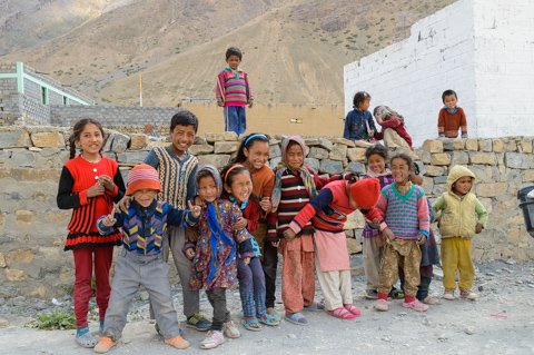 A group of smiling schoolchildren gather and pose together near Dhankar in the Spiti Valley, wearing colorful clothes against a backdrop of stone walls and barren mountains.