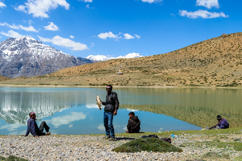 Four men sit and stand along the shore of Dhankar Lake, handlining fish beside still water that reflects snow-capped mountains in the Spiti Valley.