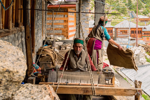 A Himalayan rug maker works at a traditional wooden loom in Chitkul, with stone walls, woven textiles, and village buildings surrounding the workspace.