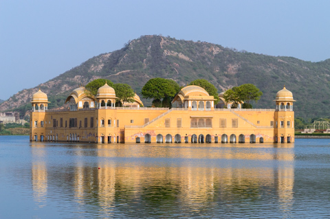 Jal Mahal palace stands in the middle of Man Sagar Lake in Jaipur, Rajasthan, India, its yellow sandstone façade reflected in the calm water with low hills in the background.