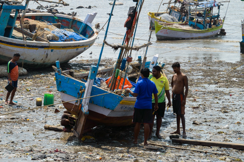 Fishermen stand beside a small wooden fishing boat along a polluted shoreline in Mumbai, India, with other boats resting in the shallow water behind them.