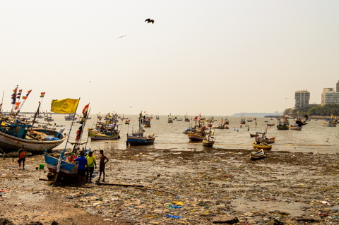 Fishing boats sit grounded along a polluted shoreline in Mumbai, India, as fishermen work among debris at low tide with the city skyline faintly visible in the distance.