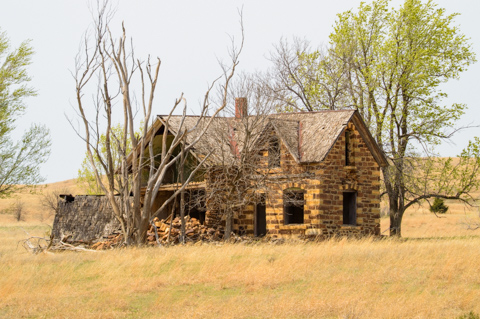The remains of an abandoned stone farmhouse stand on the open plains of Kansas, United States, surrounded by tall grass and leafless trees beneath a pale sky.