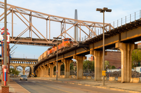 A freight train crosses an elevated rail bridge at sunrise along the riverfront in St. Louis, Missouri, with the Gateway Arch rising in the background.